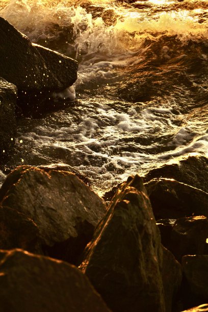夕阳拍击着海岸的岩石。金色的浪花打在岩石上。海上的日落。美丽的金色浪花腾空而起。日落时分，海浪拍打着岩石。图片下载