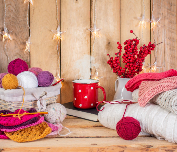一个红色的杯子，里面有热饮，篮子里有彩色的毛线球，还有奶奶方巾，钩针，灯和羊毛毯子，前面是浅棕色的木墙背景图片下载