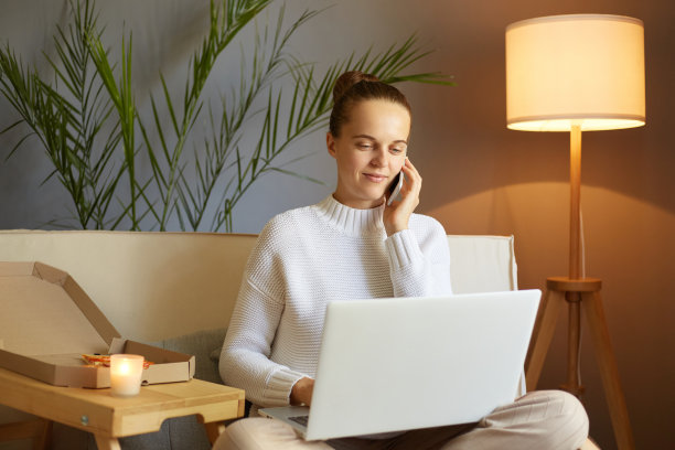 忙碌的女性肖像，深色头发，穿着白色套头衫，坐在沙发上用笔记本电脑，在键盘上打字，与她的老板或商业伙伴打电话。图片下载