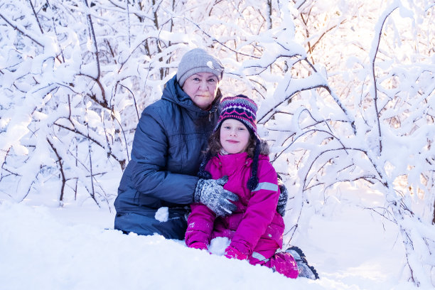肖像。一位77岁的老妇人和一个7岁的女孩微笑着看着镜头，背景是白雪覆盖的树枝和阳光。图片下载