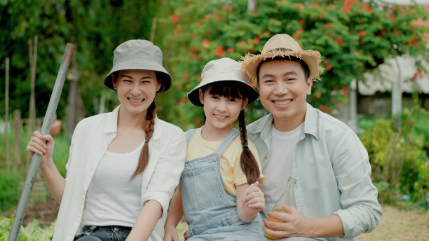 幸福的亚洲家庭，美丽的母亲，父亲和他们的小女儿微笑着看着镜头。他们戴着帽子。妈妈拿着铲子，爸爸拿着一瓶新鲜的果汁在家里的花园里。本土生产。在家种地图片下载