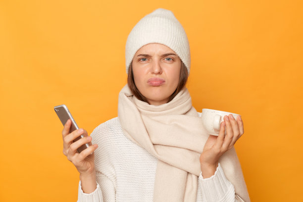 照片中悲伤的年轻女子穿着温暖的毛衣，帽子和围巾摆出孤立的黄色背景，站着，表达悲伤和悲伤，拿着杯子和智能手机。图片下载