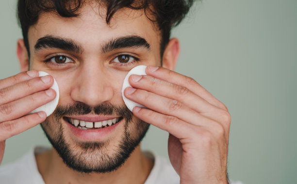 特写肖像的一个有吸引力的微笑男子手持化妆棉在他的脸颊上应用乳液孤立在粉彩背景工作室。皮肤护理保健美容程序概念图片下载