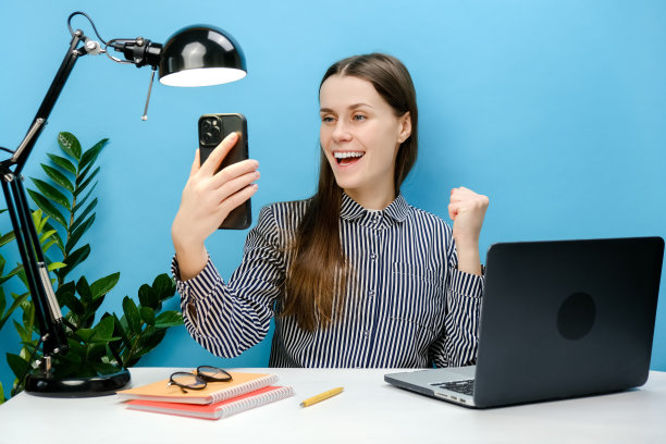 画像成功的员工年轻的商业女性在衬衫坐工作在白色的办公桌与笔记本电脑手持手机做赢家手势握紧拳头，在蓝色的工作室背景上摆出孤立的姿势图片下载