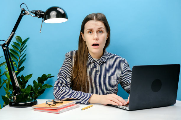 震惊的年轻员工女商人20岁在办公室坐在办公桌上的笔记本电脑电脑工作，看着相机，在工作室的蓝色背景墙外摆出孤立的姿势。事业成就图片下载