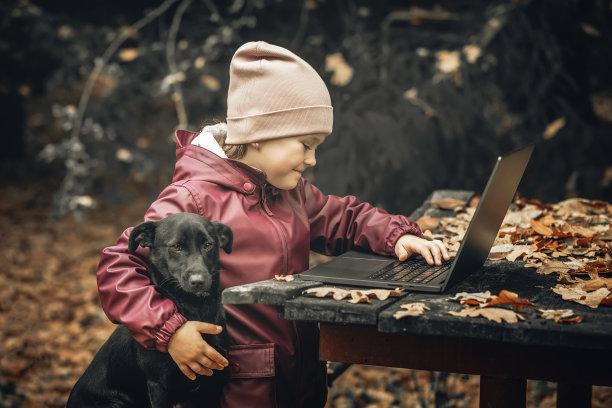 小女孩和她的狗坐在森林中央的一张旧木桌旁，用笔记本电脑进行在线学习或交流图片下载