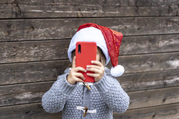 小女孩戴着圣诞老人的帽子，在圣诞节期间在户外玩一个红色的智能手机-小女孩站在一个木制背景-电子商务和圣诞礼物的技术概念图片下载