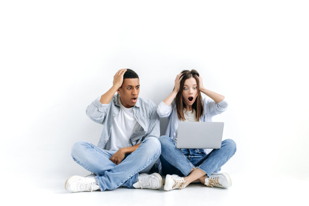 震惊气馁的年轻西班牙男子和白人女子，穿着时髦，坐在地板上孤立的白色背景与笔记本电脑，惊讶地看着屏幕，看到意想不到的消息图片下载