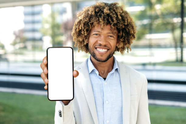 模型的概念。快乐自信的拉丁或巴西卷发男子，站在户外，展示智能手机与空白的白色模型屏幕与广告空间，演示，看着相机，微笑图片下载