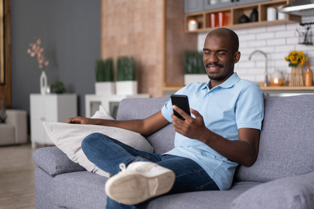 远程通信。非裔美国年轻人，看着他的手机，玩得很开心，坐在厨房房间内部的沙发上。快乐的黑人男子通过手机视频通话，呆在家里图片下载