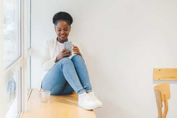 一名非裔美国妇女坐在咖啡店里喝着奶昔，靠在窗户边用手机。用手机，在她的社交网络账户上查看新闻。图片下载