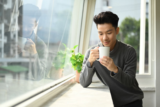 英俊的亚洲男人享受他的早上咖啡，站在办公室窗户俯瞰城市景观。图片下载