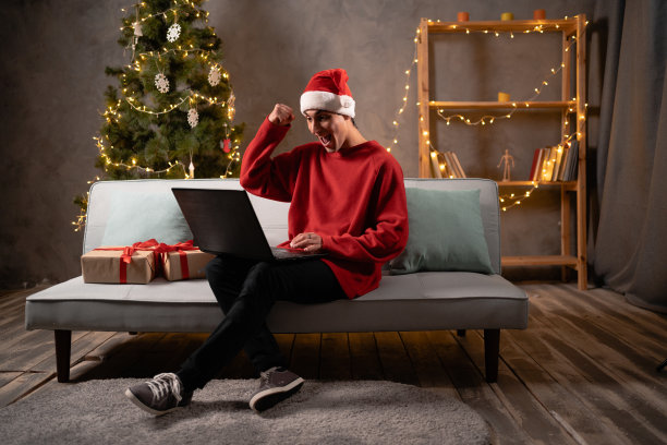 快乐的年轻阿拉伯男子在节日红帽子抱着电脑在大腿上，笑着看有趣的圣诞电影在线，休息在舒适的沙发节日庆祝图片下载