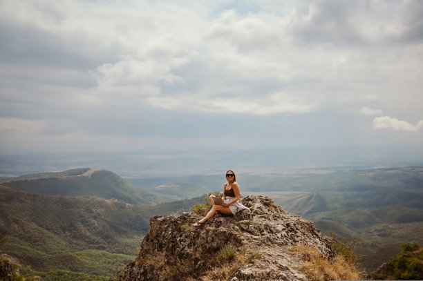 年轻女性自由职业者旅行者在户外任何地方使用笔记本电脑享受山峰的景色。快乐的女性带着太阳镜在不寻常的工作场所在夏天拿着电脑。图片下载