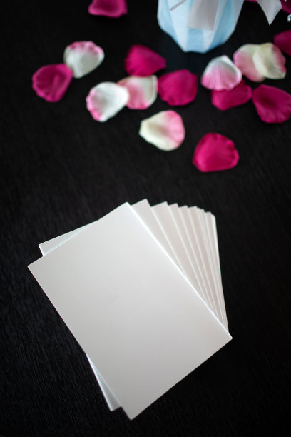 许多白色的结婚明信片放在黑色的木桌上，四周点缀着红色和粉色的玫瑰花瓣，装饰精美，显得格外出众。图片下载
