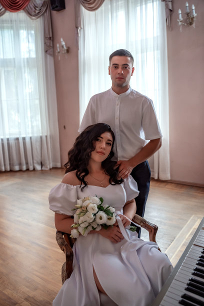 新娘手捧一束鲜花坐在婚礼大厅的钢琴前，新郎站在后面看着他爱的女人。结婚的概念。美丽的新婚夫妇图片下载