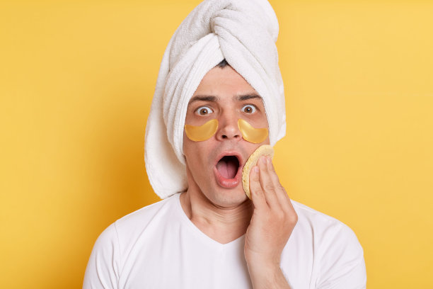 照片中，一名穿着白色t恤的男子被毛巾裹着，用海绵擦掉脸上的奶油，孤立地站在黄色背景下。图片下载
