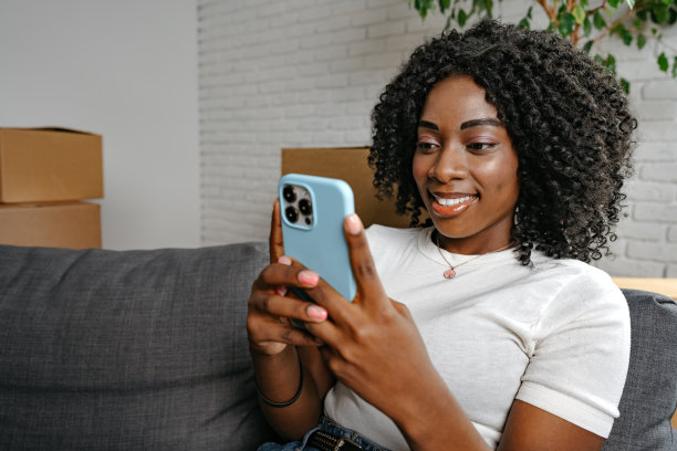 微笑的非洲女人坐在家里的沙发上用智能手机图片下载