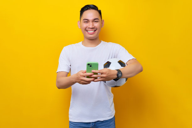 身穿白色t恤，手拿足球和黄色背景的手机微笑的年轻亚洲球迷。人们崇尚运动休闲的生活方式理念图片下载