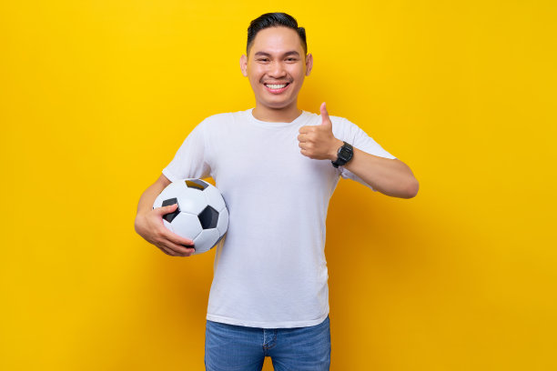 身穿白色t恤的亚洲年轻球迷拿着足球，在黄色背景下竖起大拇指。人们崇尚运动休闲的生活方式理念图片下载