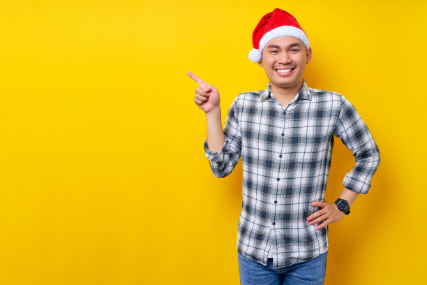 一名戴着圣诞老人帽的亚洲青年用手指指着黄色演播室背景上的广告文字空白。庆祝圣诞假期和新年的概念图片下载