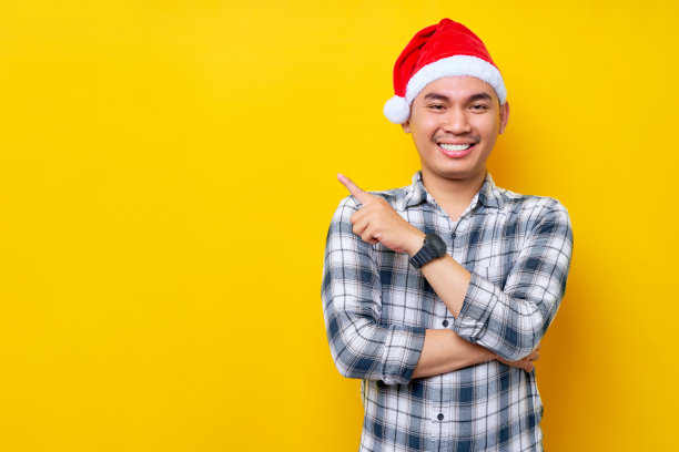 一名戴着圣诞老人帽的亚洲青年用手指指着黄色演播室背景上的广告文字空白。庆祝圣诞假期和新年的概念图片下载