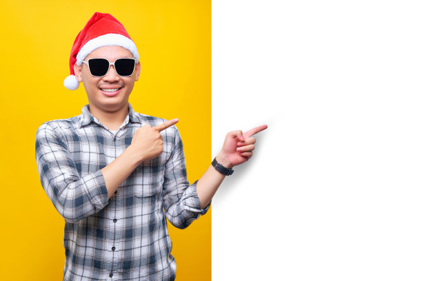 兴高采烈的年轻亚裔男子戴着圣诞帽和眼镜，指着一面孤立在黄色背景上的白色广告牌。2023年新年快乐假期概念图片下载