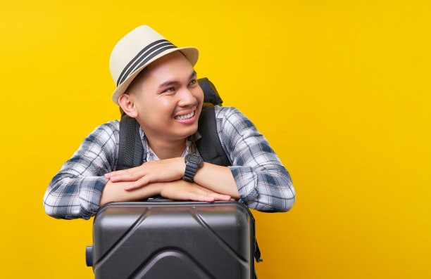 微笑的旅行者游客亚洲男子在休闲服帽子背包靠在灰色行李箱手提箱看一边在黄色背景。旅客出国旅游周末度假航空旅行概念图片下载
