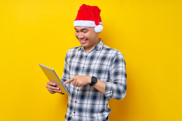 年轻英俊的微笑的亚洲人戴着圣诞帽使用和寻找宣传片上的数字平板隔离在黄色背景。圣诞节和2023年新年快乐假期概念图片下载