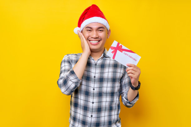 兴奋的年轻亚洲男子20多岁，戴着圣诞老人的帽子，拿着商店的礼券券卡，抚摸着孤立在黄色工作室背景上的脸颊。圣诞快乐概念图片下载