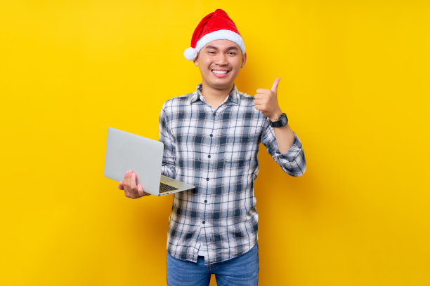 快乐的年轻亚洲男子在格子衬衫，戴着圣诞帽摆姿势站在使用笔记本电脑电脑和显示大拇指在黄色背景标志。圣诞节和2023年新年快乐假期概念图片下载