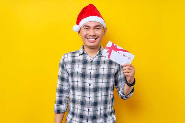 笑容开朗的年轻亚洲男子20多岁，戴着圣诞老人的帽子，拿着商店的礼券券卡，看着相机孤立在黄色的工作室背景上。圣诞快乐概念图片下载