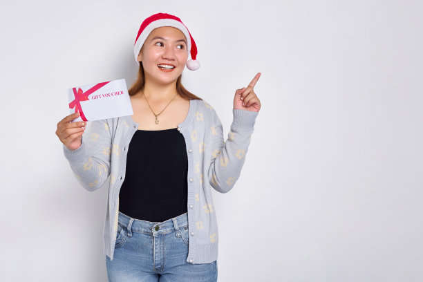 微笑年轻的亚洲女子20岁在帽子圣诞节收到礼券券券卡，指着空白空间孤立的白色工作室背景。圣诞快乐概念图片下载