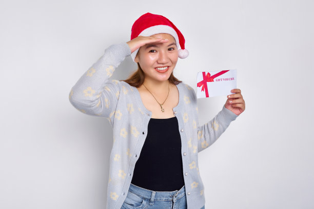 兴奋的年轻亚洲女子20多岁，戴着圣诞老人的帽子，拿着礼券券卡，看着远处孤立的白色工作室背景。圣诞快乐概念图片下载