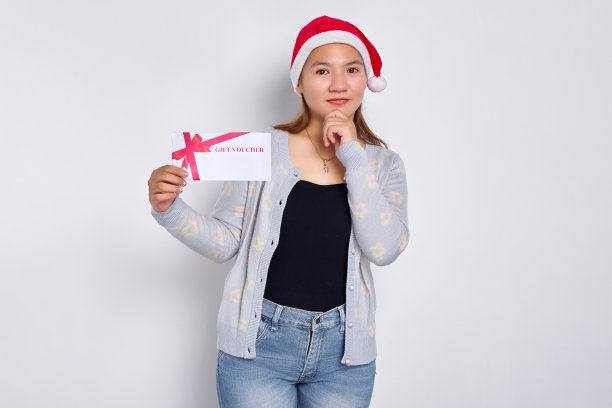 思考和沉思的年轻亚洲妇女在圣诞老人的帽子拿着礼券券卡，触摸她的下巴与手孤立在白色的工作室背景。圣诞快乐概念图片下载