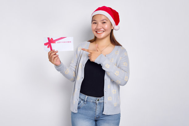 微笑年轻的亚洲女子20岁在帽子圣诞节指着一个礼券券券卡孤立在白色的工作室背景。圣诞快乐概念图片下载
