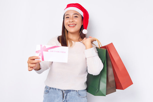 微笑的年轻金发亚洲女子20岁戴着圣诞帽，手持礼券证书和购物袋在白色背景上孤立。2023年新年快乐假期概念图片下载
