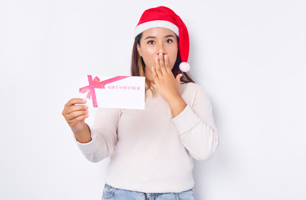 震惊害羞的年轻亚洲女子20岁在帽子圣诞节显示礼券券券卡，覆盖口与手孤立在白色工作室背景。圣诞快乐概念图片下载