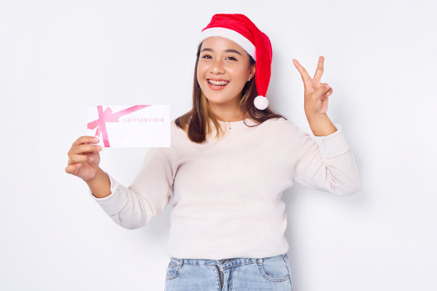 自信的年轻亚洲女子20岁在圣诞老人的帽子显示礼券券券卡，显示胜利v手势在白色工作室背景。圣诞快乐概念图片下载