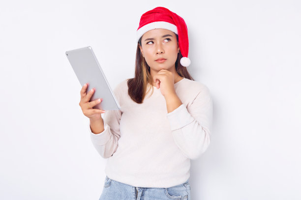 一名戴着帽子的沉思的亚洲年轻女子用平板电脑过圣诞节，抚摸着白色背景下孤立的脸颊。印尼人庆祝圣诞节的概念图片下载
