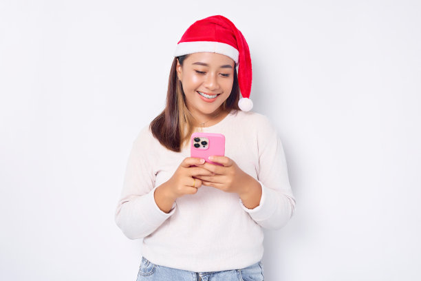 戴着圣诞帽的微笑年轻亚洲女子看着智能手机，在白色背景下获得了好消息。庆祝圣诞假期和新年的概念图片下载