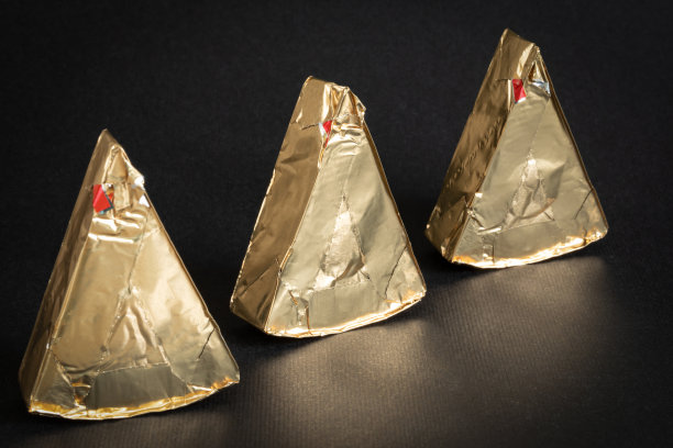 三块三角形的融化的奶油奶酪用金色铝箔纸包裹在黑色背景上。图片下载