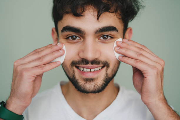 一个微笑的英俊男子清洁面部皮肤与两个棉垫在灰色背景看着相机的正面视图肖像。孤立的肖像。美容治疗。皮肤护理。皮肤病、美容。美丽的脸。图片下载