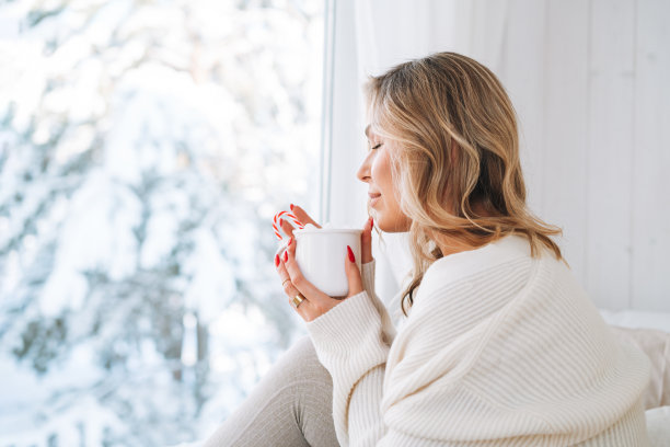 年轻女子在白色开衫与一杯可可看着窗口与冬季景观在家里图片下载