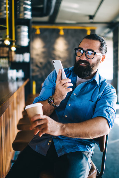 微笑的成熟白人男子在咖啡休息时间用手机聊天和写博客，快乐的40岁大胡子男子拿着连接4G网络的智能手机在室内浏览图片下载