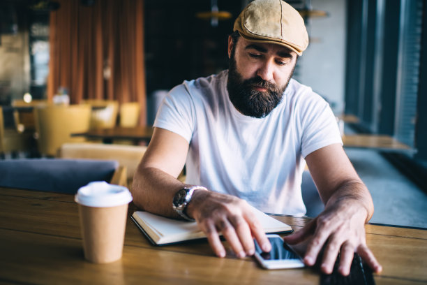 严肃的大胡子男性在咖啡馆内部工作，在笔记本上写作，在智能手机上进行研究，戴着时尚帽子的沉思的成熟男性通过连接4G的智能手机上的应用程序进行预订图片下载
