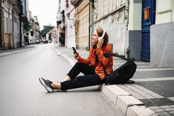 一名女子坐在路边用耳机和手机听音乐。图片下载