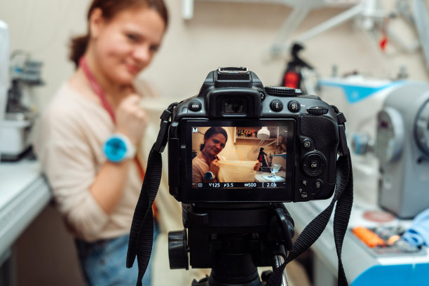 一名白人女裁缝在家庭作坊的缝纫机上缝纫，并用摄像机记录下广播。小生意。博客的概念。图片下载