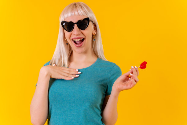 微笑着吃心形棒棒糖，金发白人女孩在工作室的黄色背景图片下载