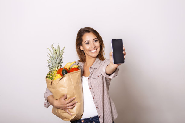 年轻女子拿着装满杂货的纸袋，展示她的手机，网上订购的概念。购物应用程序图片下载